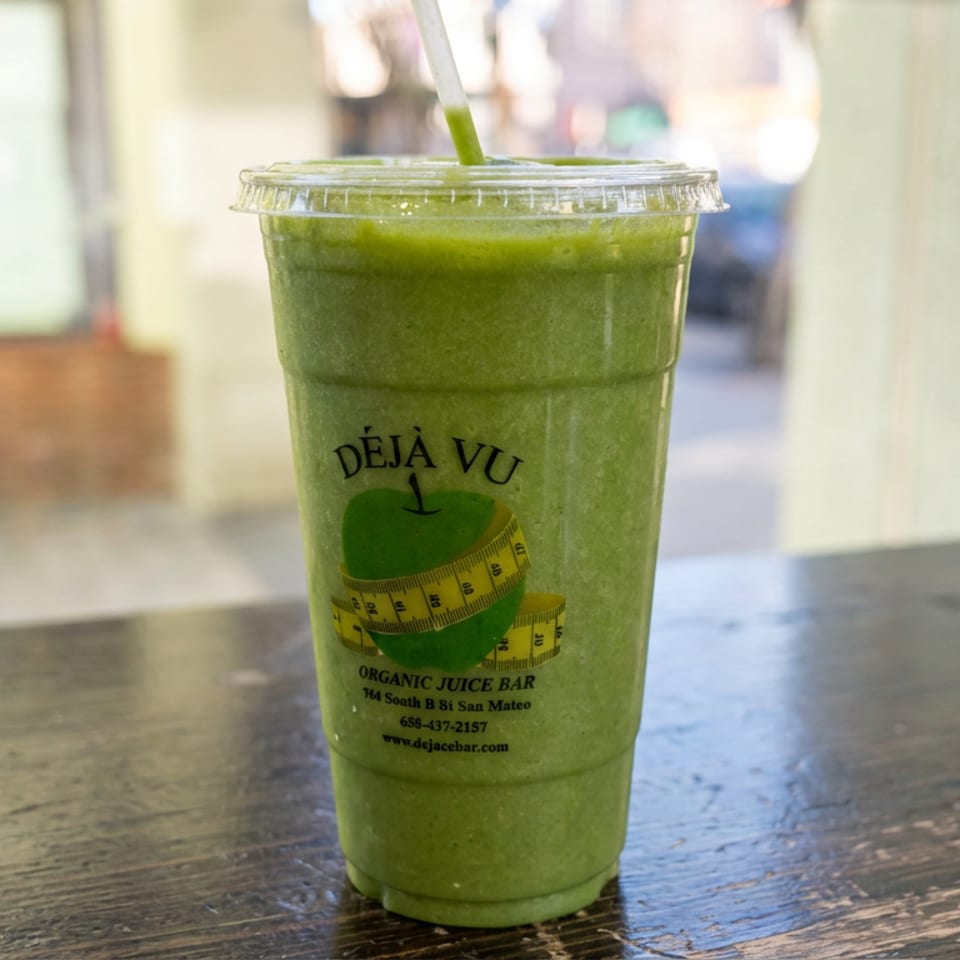 Green smoothie in a clear cup with a straw, branded Déjà Vu Organic Juice Bar, sits on a wooden table.