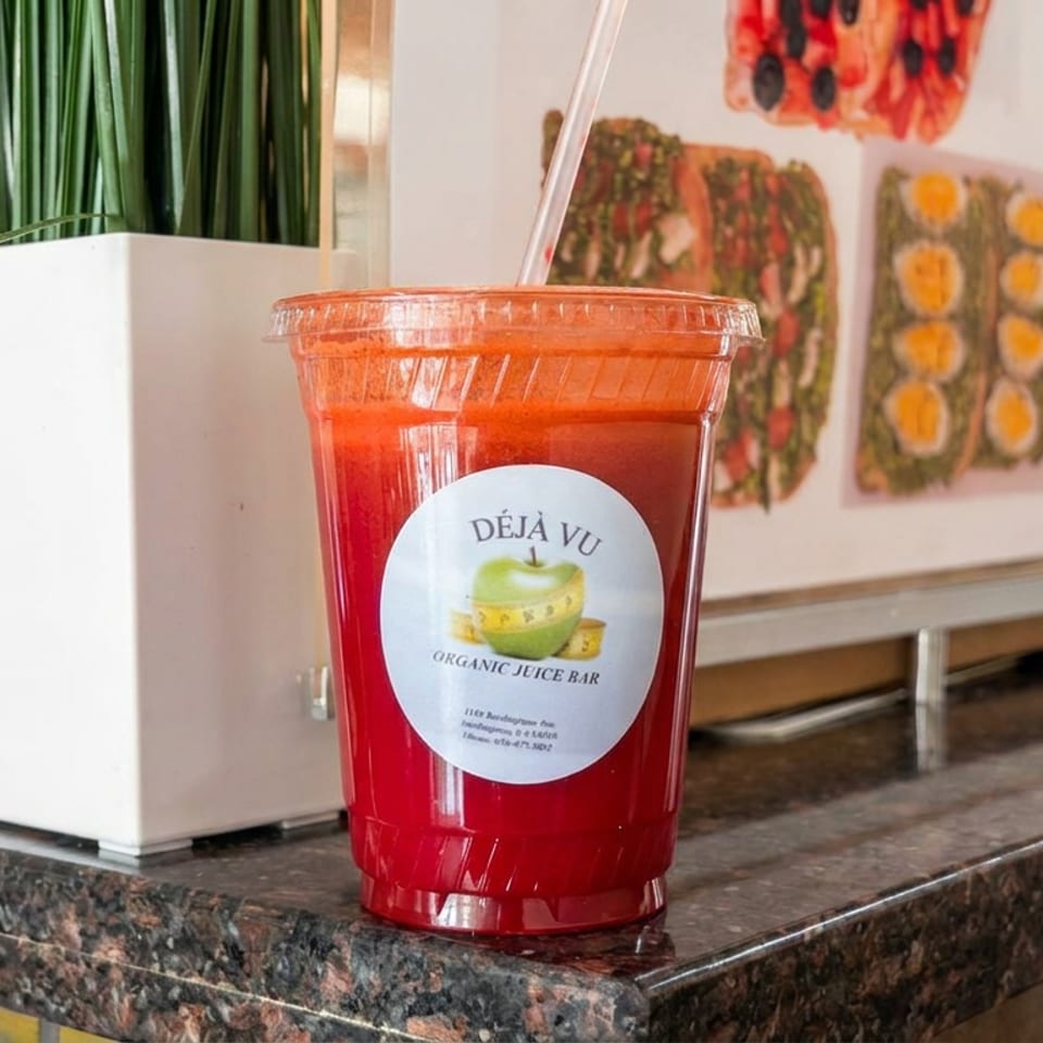 Red juice in a clear cup labeled Déjà Vu Organic Juice Bar sits on a countertop beside a planter and food artwork.