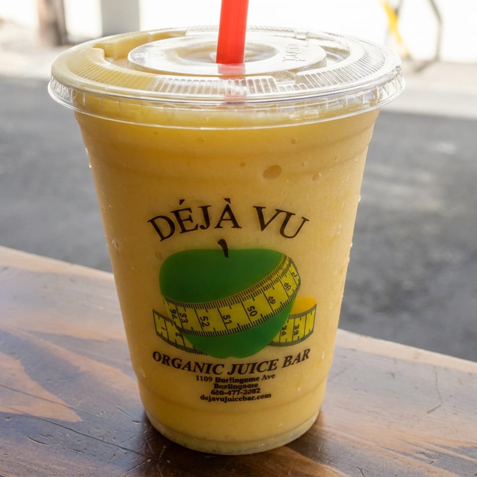 Yellow-orange smoothie in a clear lidded cup with a red straw, branded Déjà Vu Organic Juice Bar, sitting on a wooden table.