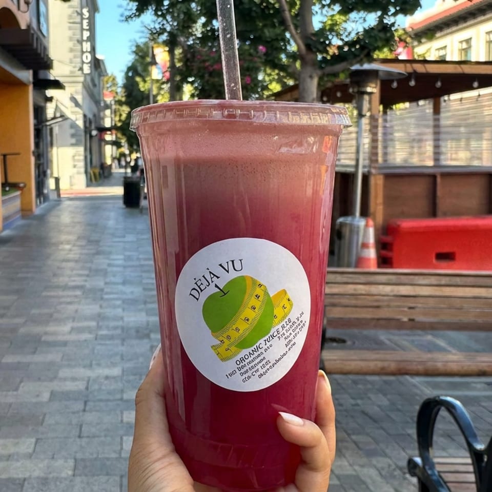 Hand holding a red organic juice smoothie labeled Déjà Vu on a sunny urban sidewalk.