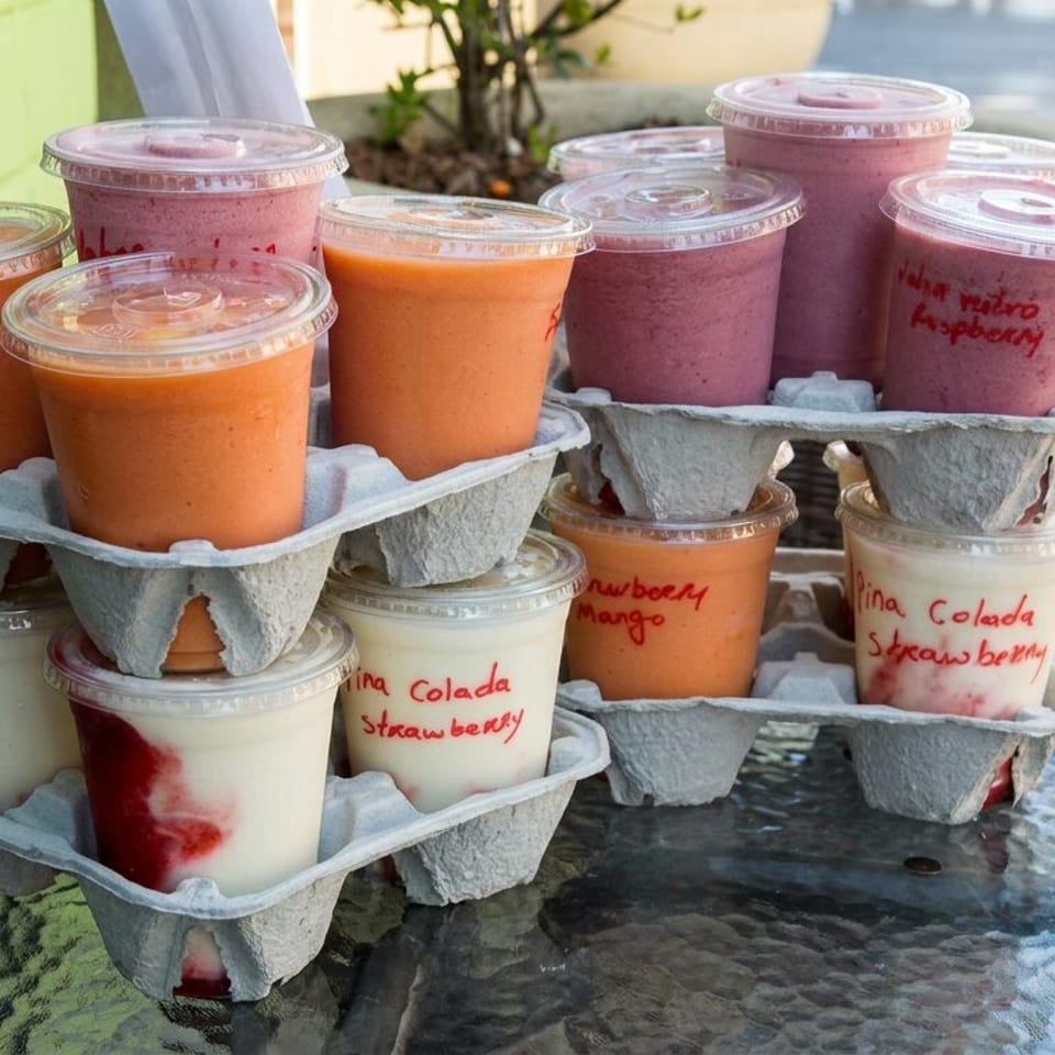 Colorful fruit smoothies in labeled plastic cups arranged in cardboard carriers on an outdoor café table.
