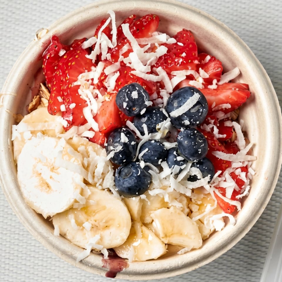 Fruit bowl topped with sliced strawberries, blueberries, bananas, and shredded coconut beside a white spoon.